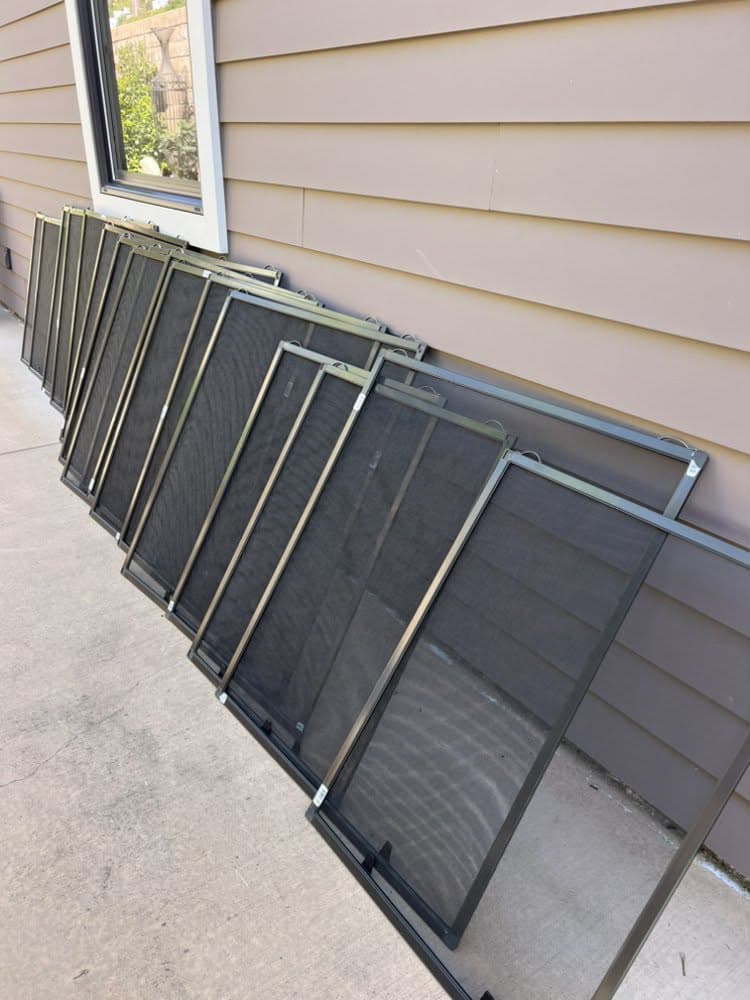 A row of new dark aluminum-frame window screens leaning along the side of a home, made by Screenova Solutions.