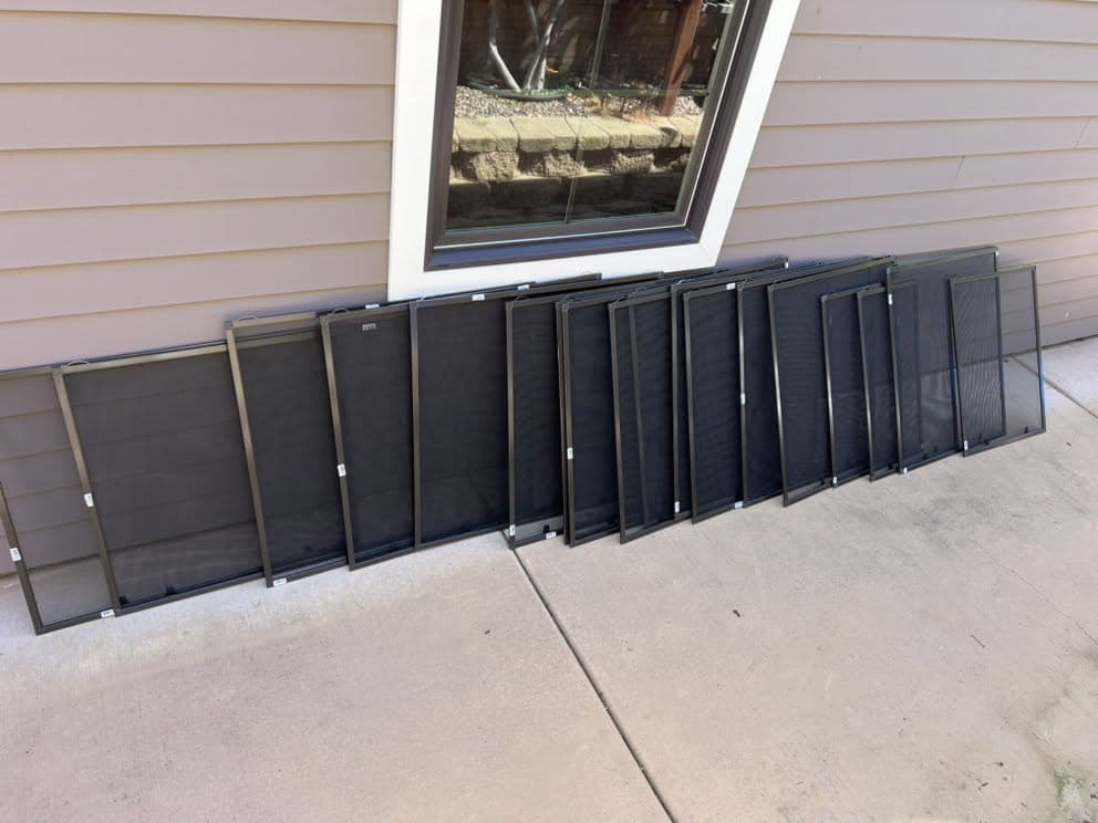 A full set of dark aluminum-frame window screens with dark mesh lined up beneath a window, ready for installation.