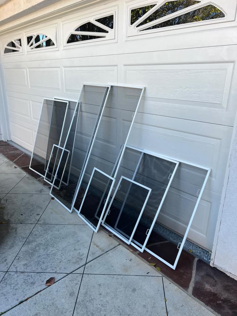 Seven custom white window screens in assorted sizes leaning against a white garage door, ready for installation by Screenova Solutions.