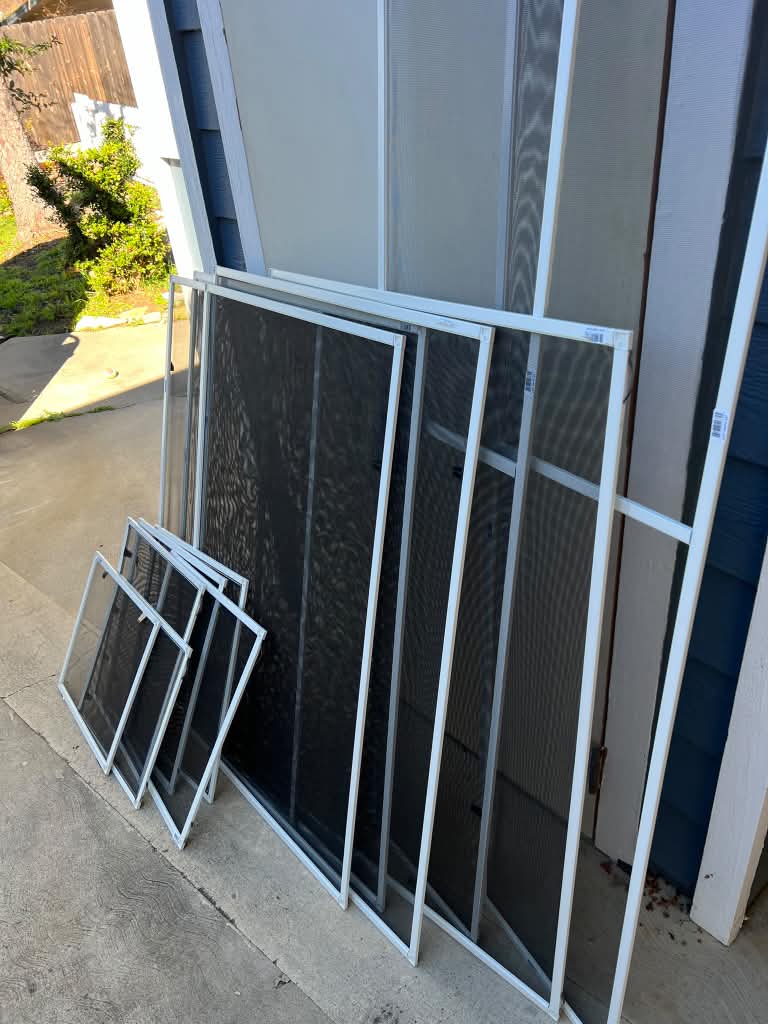 A close-up of brand-new white window screens with fresh mesh leaning against a blue house.