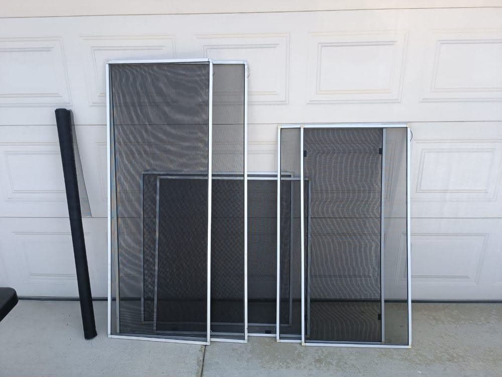 Silver aluminum-frame window screens beside a roll of charcoal screen mesh against a white garage door.