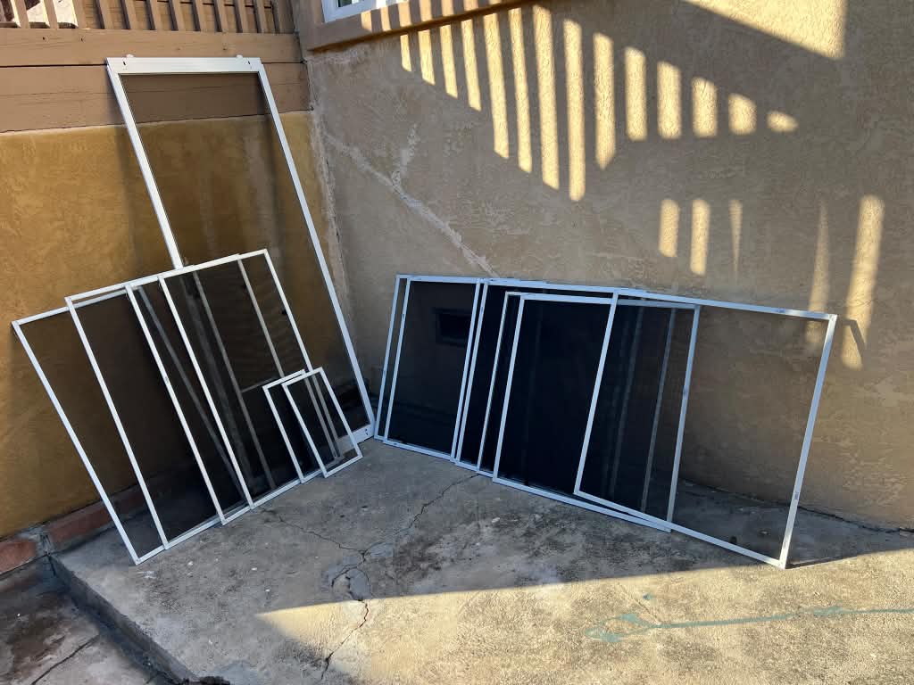 New window screens and a tall door screen staged in the corner of a sunny patio before installation by Screenova Solutions.