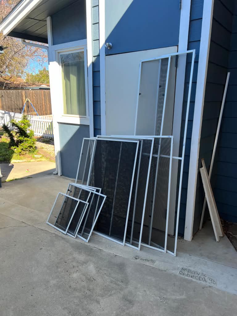 Custom white window screens and a tall screen ready for installation outside a blue-sided home, by Screenova Solutions.