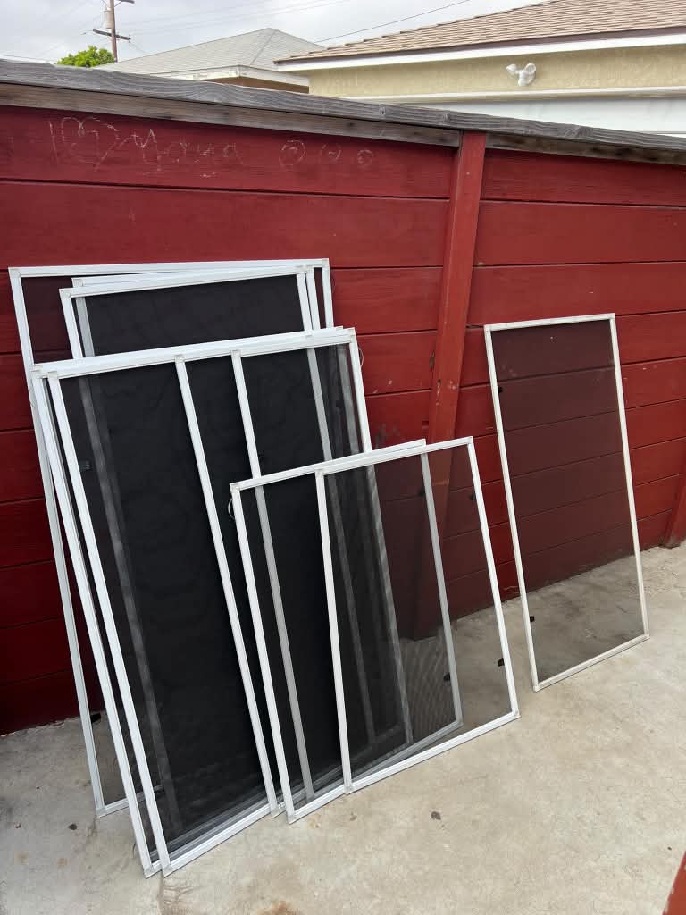 New custom window screens in multiple sizes leaning against a red wooden fence before installation.
