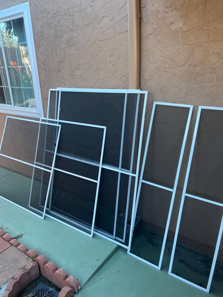 Custom window screens with horizontal crossbar supports leaning against a tan stucco home by Screenova Solutions.